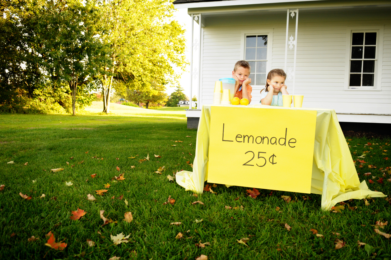 A young boy and girl stand bored behind their lemonade stand.